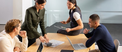 A group of people sitting in a meeting with a laptop on a table and one person looking at a tablet.