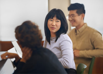 Three people are seated indoors, engaged in conversation, with laptops open in front of them.