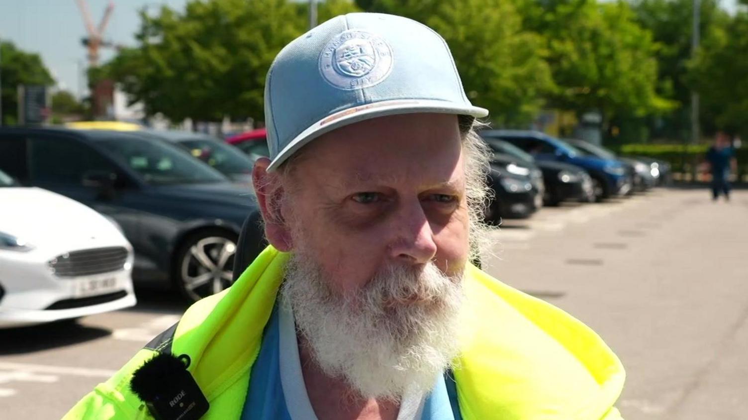 Brian Mitchell has white hair and white beard. He wears sky blue branded Manchester City baseball cap and yellow high-visibility anorak. There are cars behind him. He is the focus on the photo.