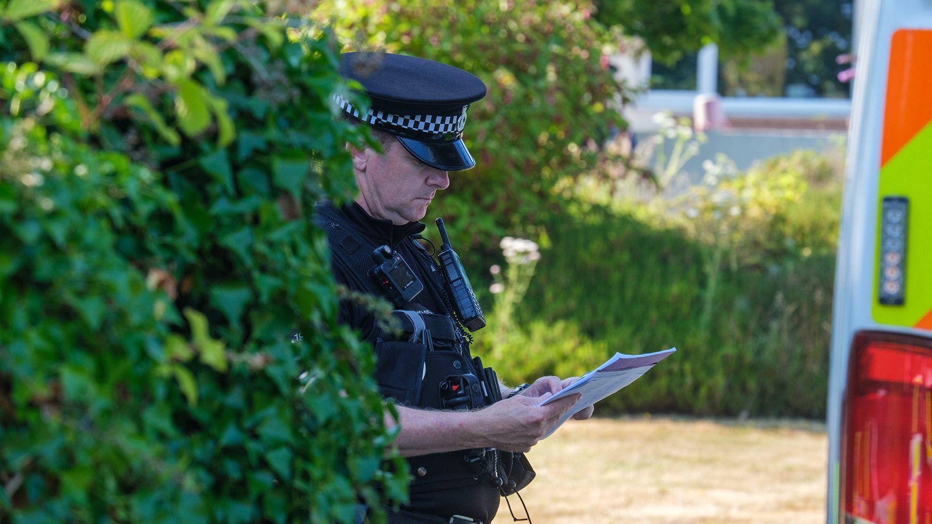 A police officer looks down at a sheet of paper while wearing uniform next to a bush and police van outside Widdecombe's home on Friday.
