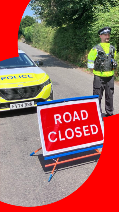 A police officer stood next to a police car and a 'road closed' sign
