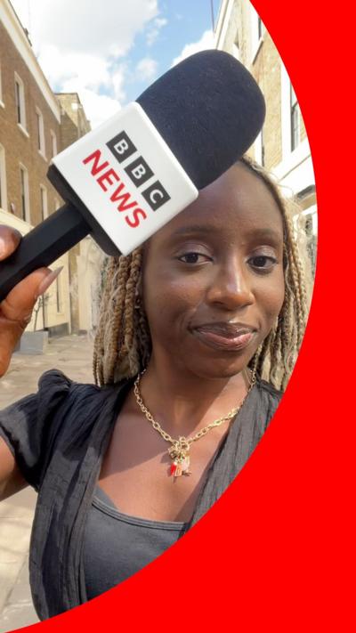 Journalist holding a BBC NEWS microphone outside on a city street.