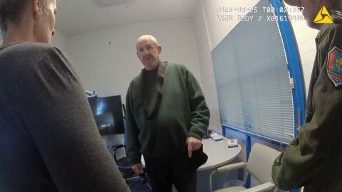 Murdoch wearing dark green jumper stands in front of camera while questioned by two officers in front of him
