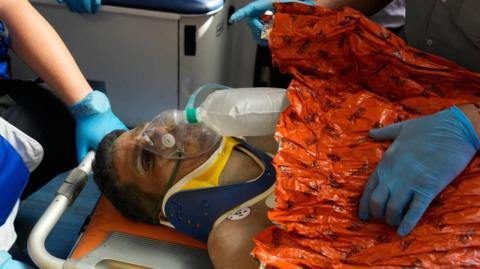 Hernán Gil is seen with an oxygen mask, a neck collar and wrapped in an orange blanket, on a stretcher