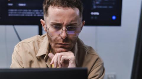 A software engineer, wearing a light brown shirt, looks at a computer monitor in front of him with a concerned expression. A larger screen with computer code on it is on a wall behind him.