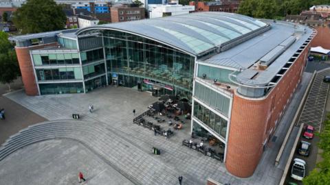 An aerial view of the BBC building at The Forum in Norfolk shows a grey roof and a large glass frontage. In front of the building is a spacious concourse paved with grey slabs, with several cars parked alongside.
