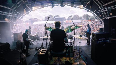 A band playing on a stage, viewed from the back, behind the drummer. In the distance is a crowd,