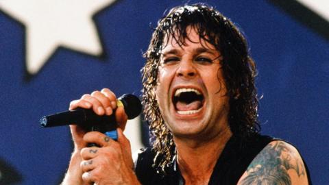 Ozzy Osbourne holds a microphone with his mouth wide open