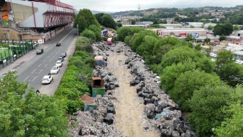 Piles of rubbish lay strewn on a long strip of land surrounded by trees. On the left hand side is a road and one side of a football stadium can be seen. To the right is an industrial area filled with low-rise factory buildings. 