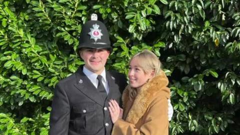 A man in police uniform stands with a blonde woman in a long brown coat.  