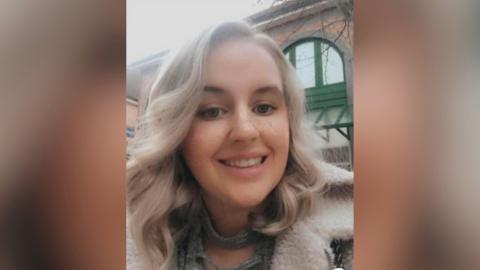 Abbigail Smith stands outside a brick building with large curved windows. She has light blonde hair down to her shoulders and is wearing a fur-lined jacket. She is smiling at the camera.
