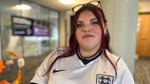 Trinity wears a white England football shirt and has glasses perched on her head. She has long brown hair. 