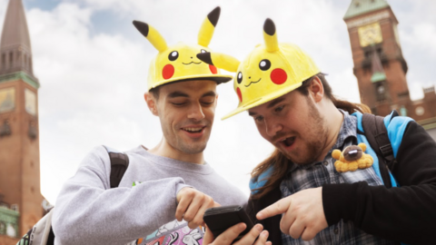 Two Pok&eacute;mon Go fans wearing hats shaped like the yellow Pok&eacute;mon character Pikachu are pointing and looking at a phone, in shock. 