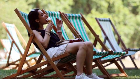 A woman sits on a deckchair the shade while eating an ice-cream at a park in London