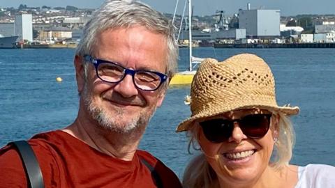 A man in a brown top with grey beard and hair with glasses and a woman in a straw hat wearing sunglasses in front of the sea.
