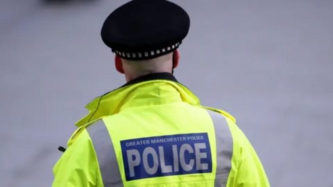 A back shot of a Greater Manchester Police officer in a green jacket and black cap