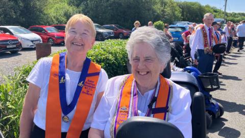 Two women enjoying the parade on a very sunny day in Rossknowlagh, County Donegal.  The woman on the left has short blonde hair and glasses and the woman on the right has short, grey hair.  They are both wearing short-sleeved white tops, orange collarettes and they are smiling broadly. 