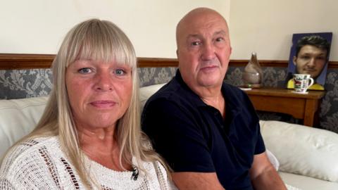 Sitting together at home on a white leather sofa, Helen Low, with long blonde hair and wearing a white top, and Martyn Low, bald and wearing a dark blue short-sleeved shirt, look at the camera lens. Behind them is a picture of their son, George.