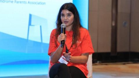 A woman with dark brown long hair and a bright orange tshirt sits on a chair on a stage holding a microphone to her mouth. She's wearing a white pass on a lanyard. Behind is a big screen with a blue background.