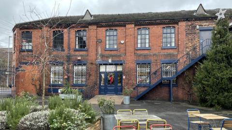 The exterior of the Emma Bridgewater gift shop which is made from red brick with windows and doors painted in blue trim. There is a Christmas wreath on the door and tables, chairs and plants can be seen in the foreground. 