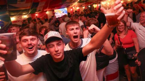 England supporters cheer after their team's first goal as they watch the television broadcast of the 2026 World Cup Group L football match between England and Panama, at The Clock pub in Hebburn, north-east England. The scene has a large crowd with three men in the front celebrating with their arms up. 