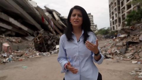 Yogita wears a blue shirt and speaks to the camera as she stands in front large piles of rubble, including what looks like a collapsed apartment block.