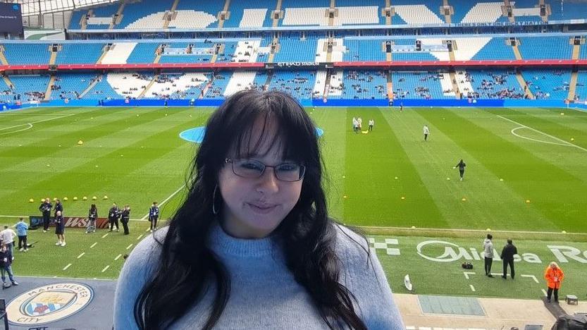Emily Brobyn has long black hair, dark spectacles and is wearing a sky blue sweater. Behind her is the pitch at Manchester City FC with the rest of the stadium in the background.