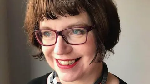 A woman with a brown bob is looking off to the side with a lovely smile. She is wearing glasses. 